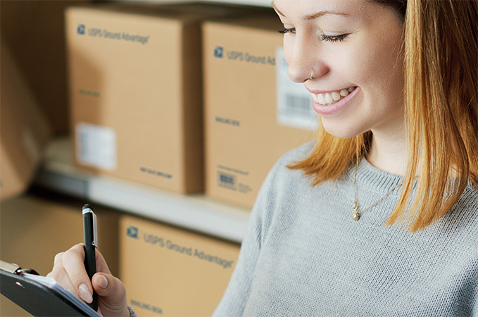 USPS Ground Advantage. A person preparing to ship packages with a shelf of USPS Ground Advantage packages behind them.